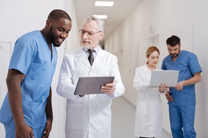two healthcare workers discussing something in the forefront while 2 other healthcare workers look at a laptop in the background of the hallway