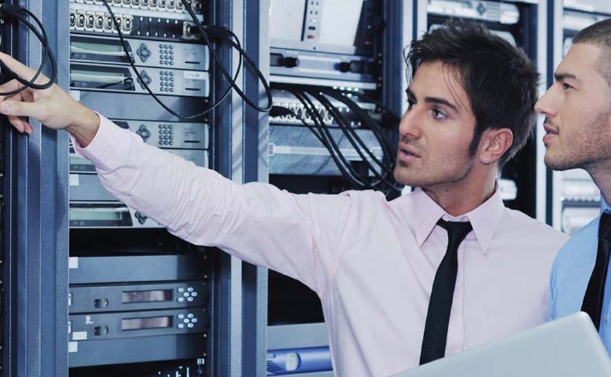 Two IT professionals working in a server room, one pointing at network cables while the other observes.