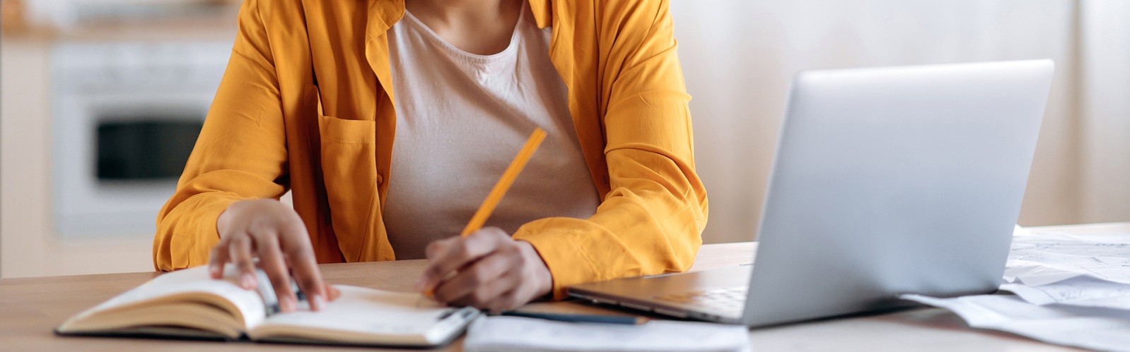 woman with braids in a bun wearing an orange shirt sitting at a desk looking at a laptop