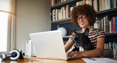 woman with brown curly hair and glasses smiling sitting at her laptop with a french bulldog on her lap