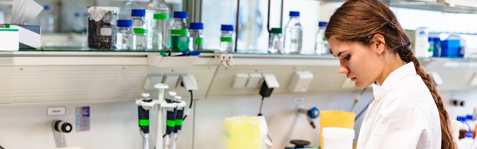 Young lady in a white lab coat with long brown hair studying a book in a pharmacy lab