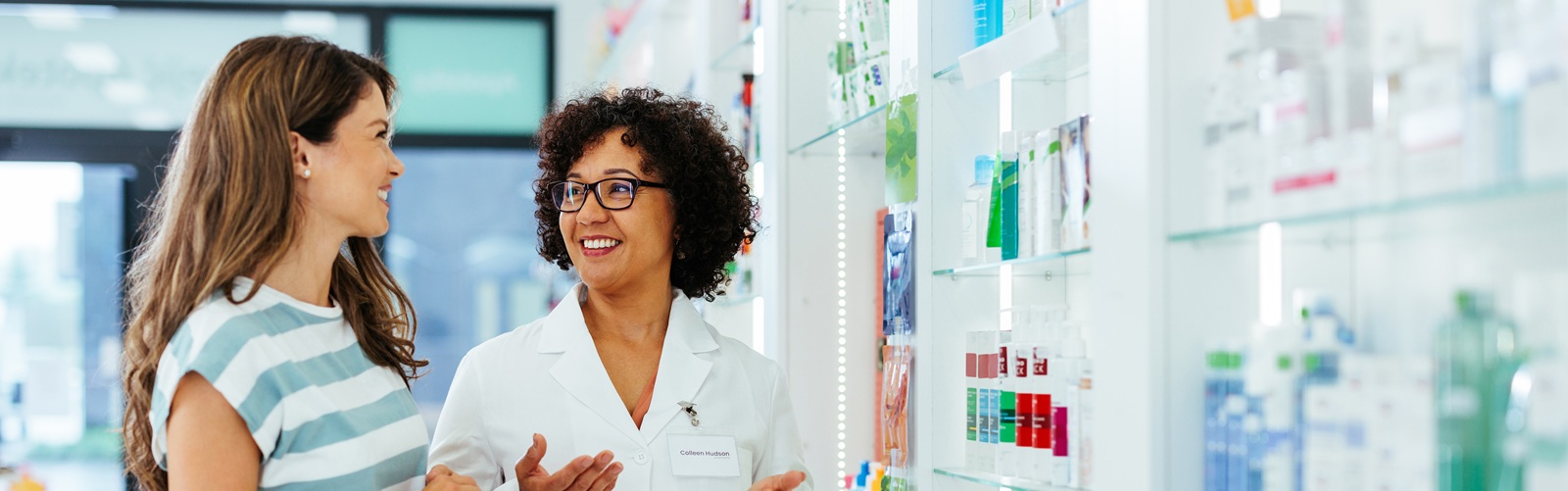 female patient speaking with a female pharmacist in a pharmacy with shelves of drugs along the wall