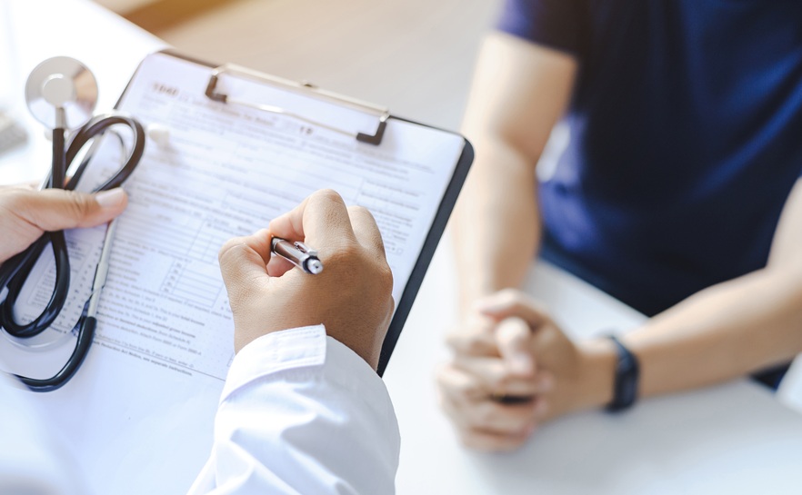 healthcare professional holding a stethiscope and a clipboard with another person across the table