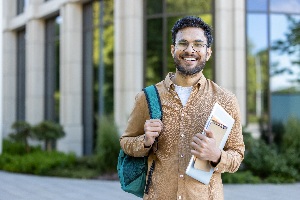 bearded man holding a backpack and book standing in front of a building