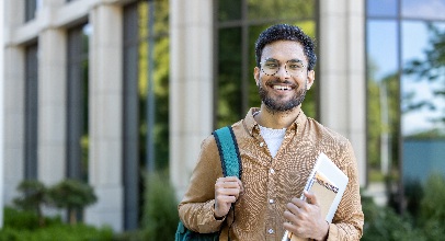 bearded man holding a backpack and book standing in front of a building