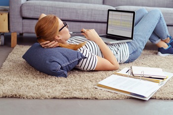 young redheaded lady laying down on the floor looking at a laptop with a notebook by her side