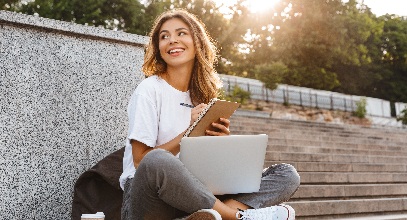 young brunette lady sitting cross legged near stairs with a laptop in her lap smiling